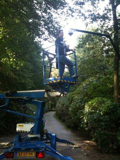 Monteur in een hoogwerker monteert een camera aan een gebogen mast boven een beschaduwde oprijlaan tussen de bomen