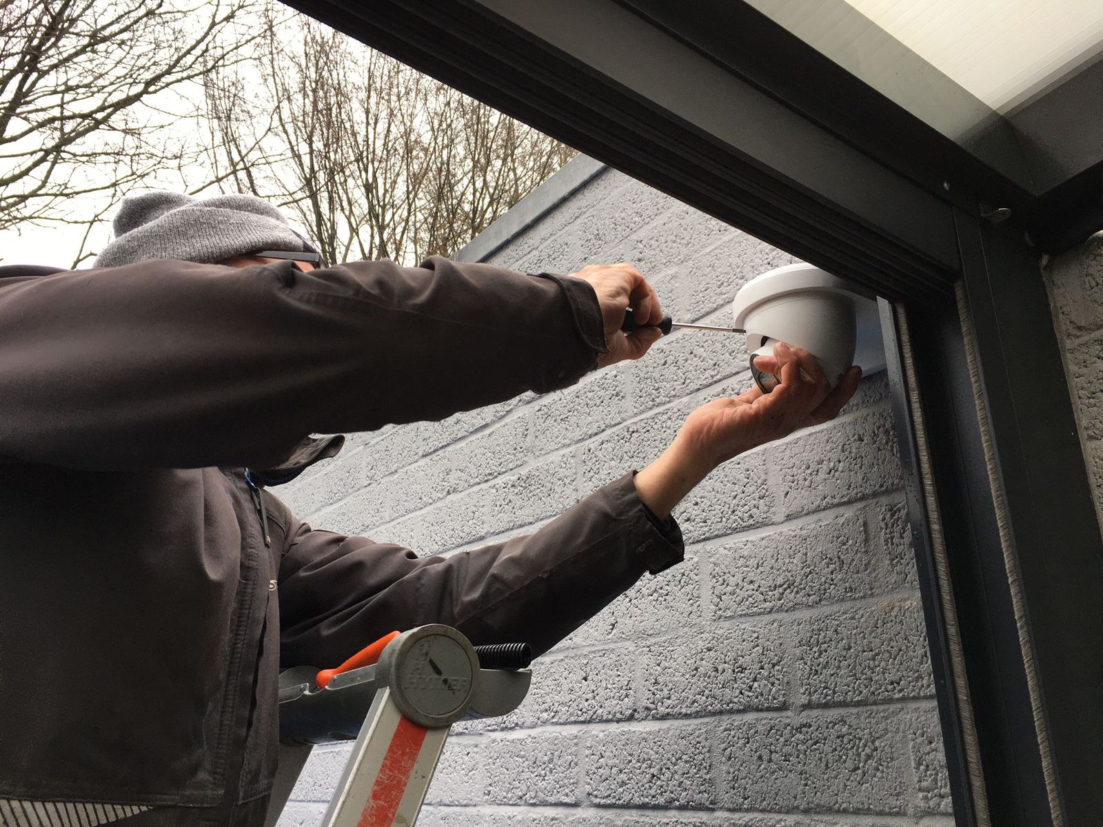 Monteur stelt een witte camera af boven de entree van een zakelijk pand met grijs metselwerk