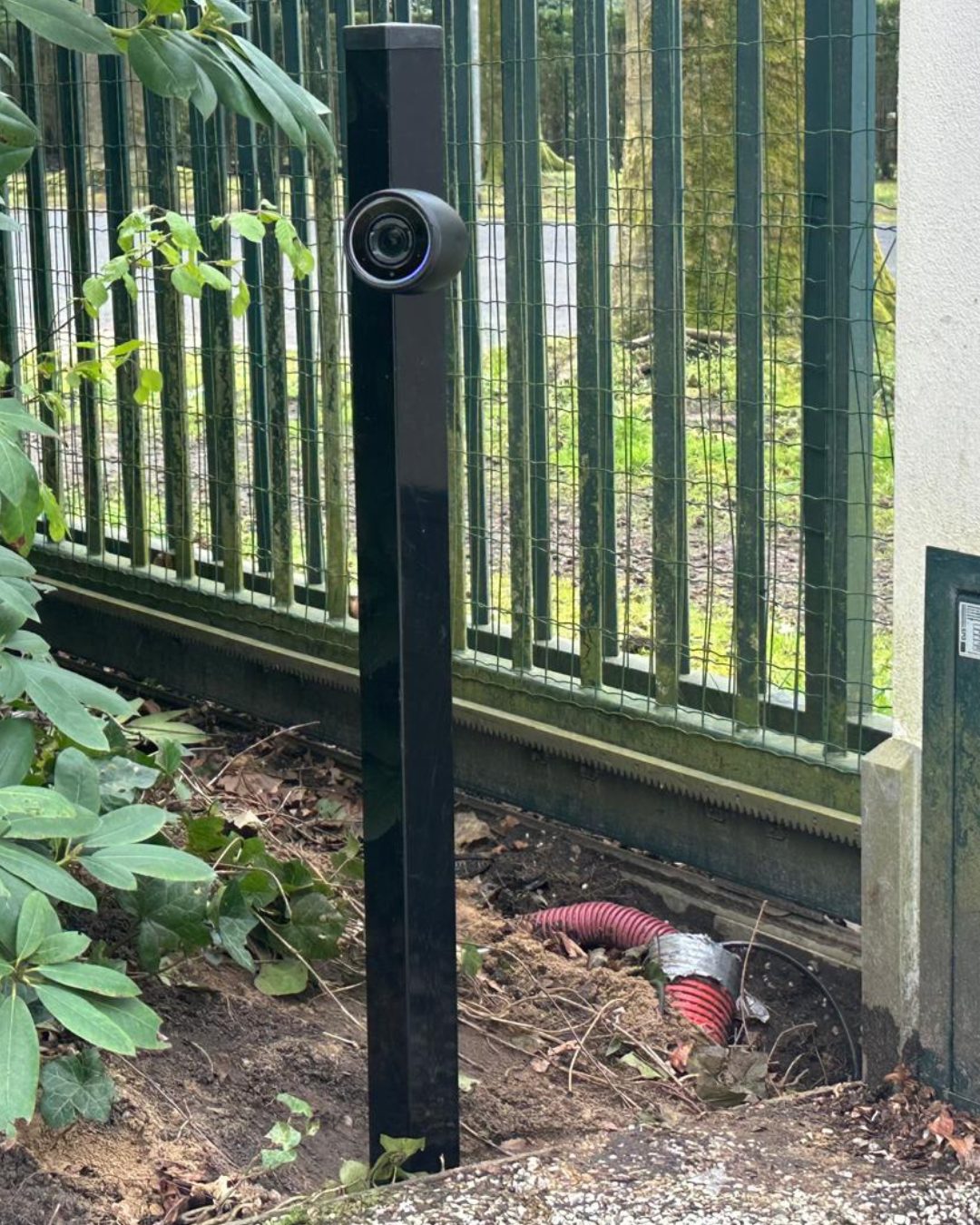 Zwarte camera met verlichte statusring op een donkere paal bij de erfgrens, de rode mantelbuis verdwijnt de grond in
