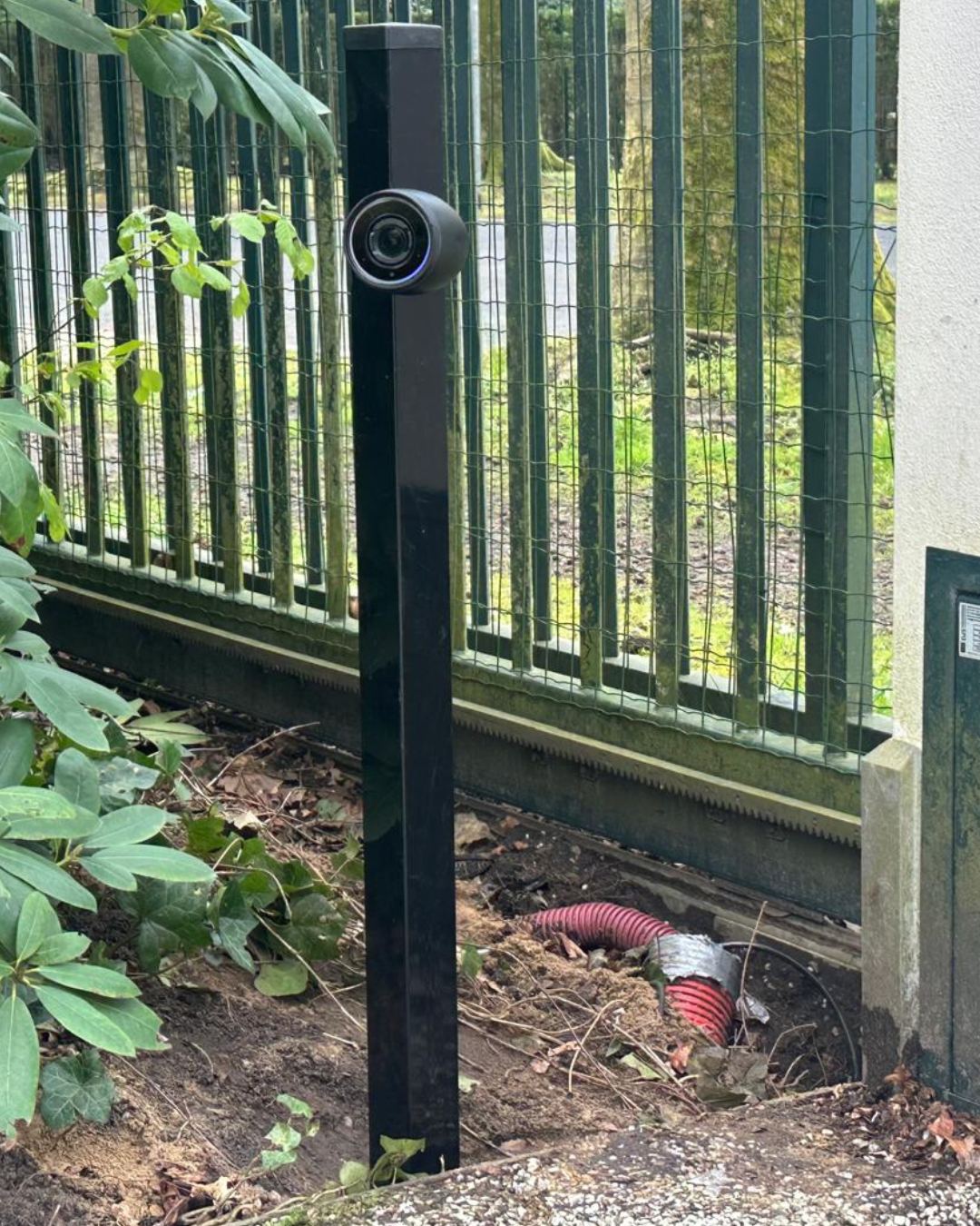 Zwarte camera op een zwarte paal bij een groen spijlenhek, met een rode mantelbuis die uit de grond komt