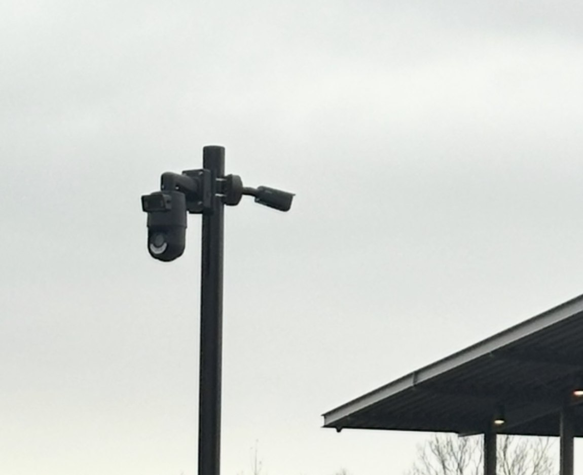 Zwarte mast met draaibare camera en vaste camera bij een overkapping op een bedrijfsterrein