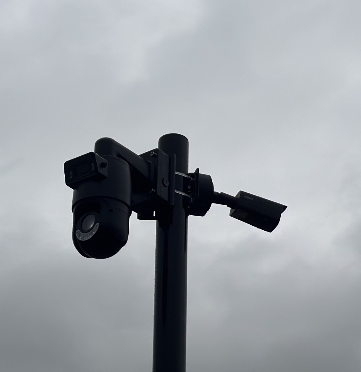 Zwarte mast met een draaibare camera en een vaste camera, als silhouet tegen een grijze wolkenlucht