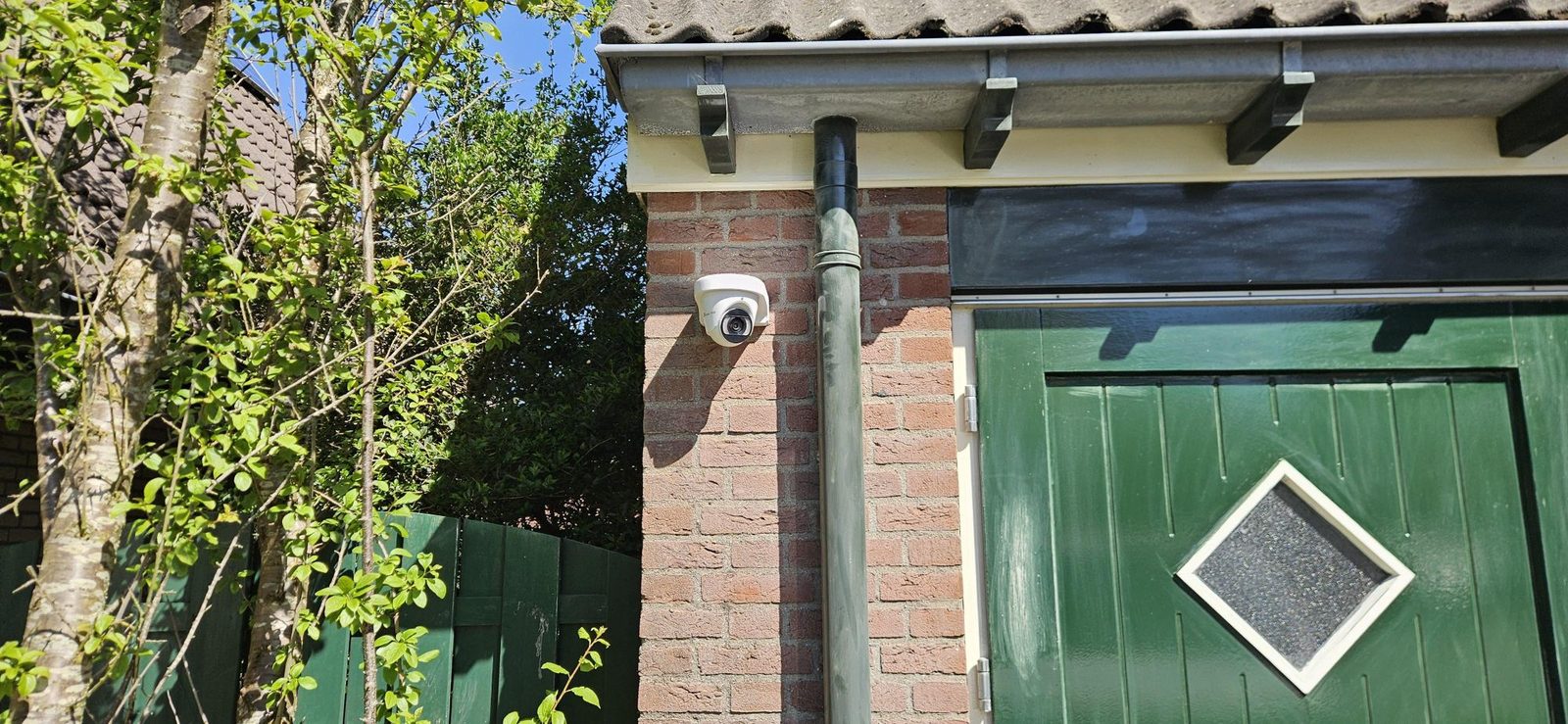 Witte camera op een gemetselde penant naast een groene garagedeur, aan een smal achterom met schutting en groen