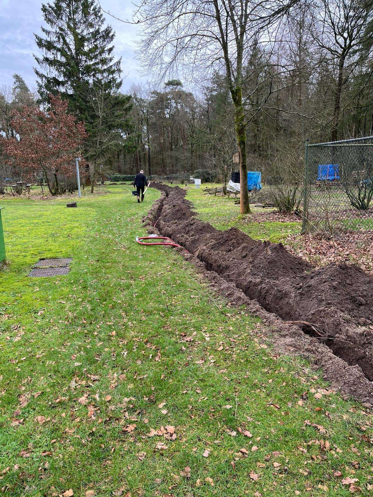 Smalle, kaarsrechte kabelsleuf door een gazon in een bosrijke tuin, met een monteur op de achtergrond