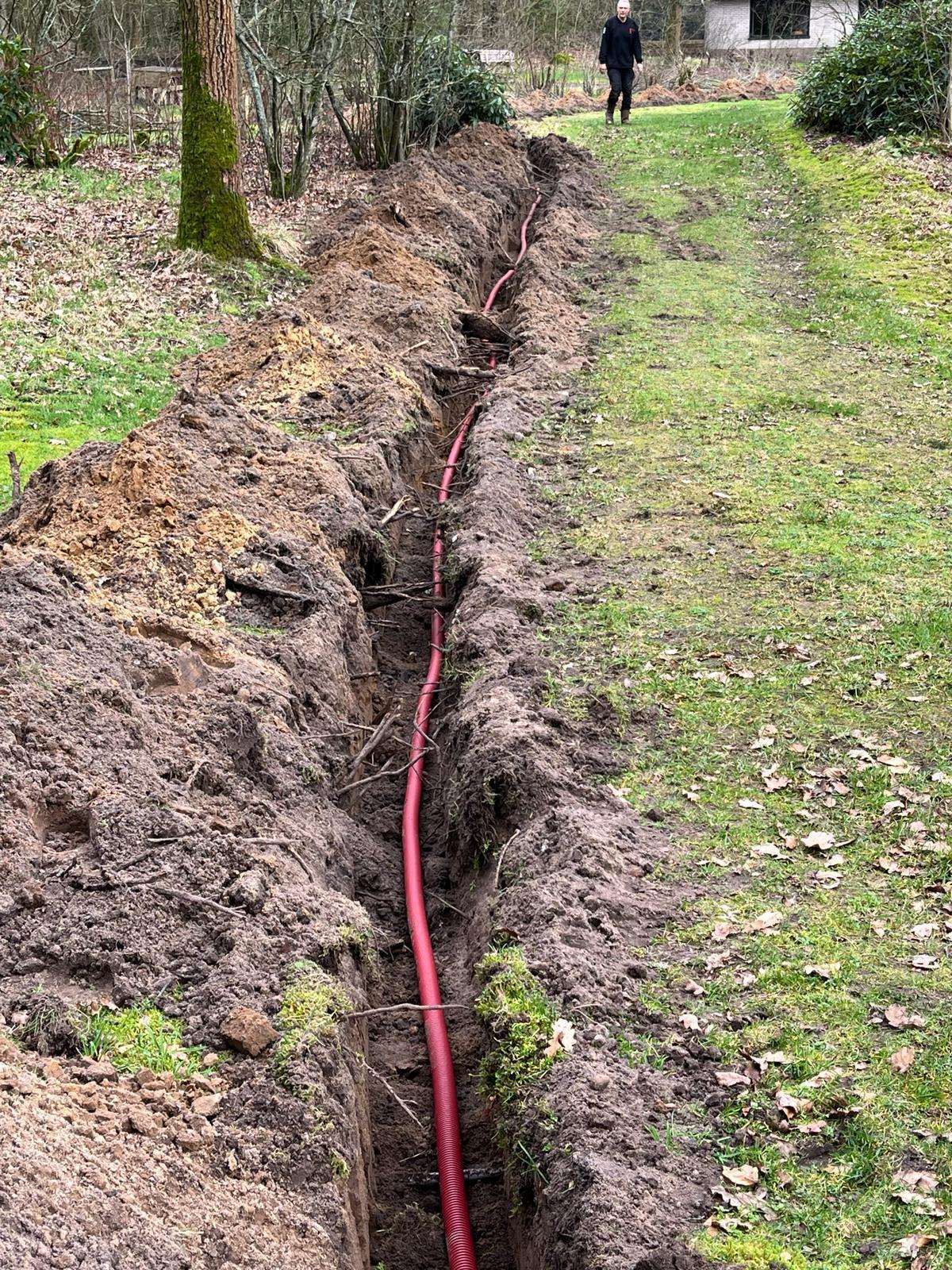 Open sleuf met rode mantelbuis die tussen bomen en struiken over een terrein naar een woning loopt