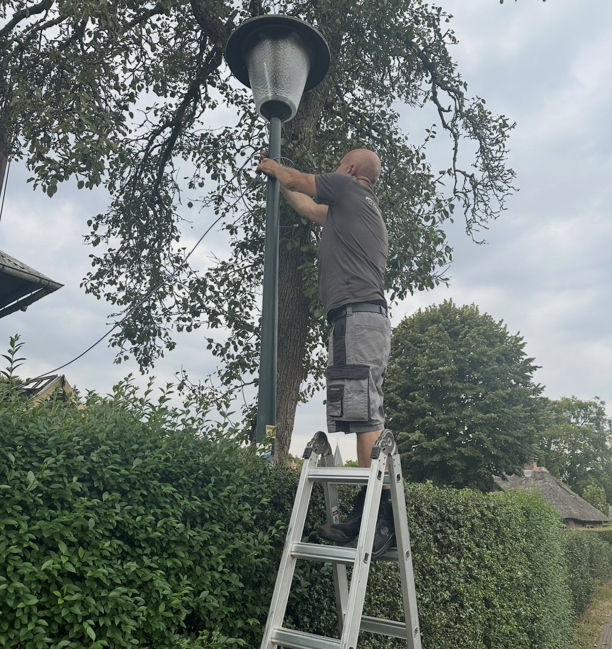 Monteur op een ladder werkt aan een klassieke dorpslantaarn tussen hoge hagen, met een kerktoren op de achtergrond