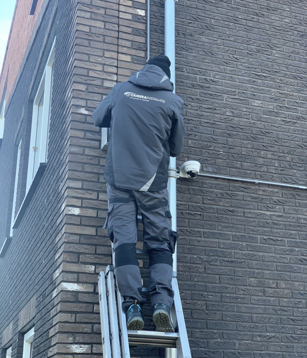 Monteur op de ladder monteert een camera strak naast de regenpijp van een donkere bakstenen gevel