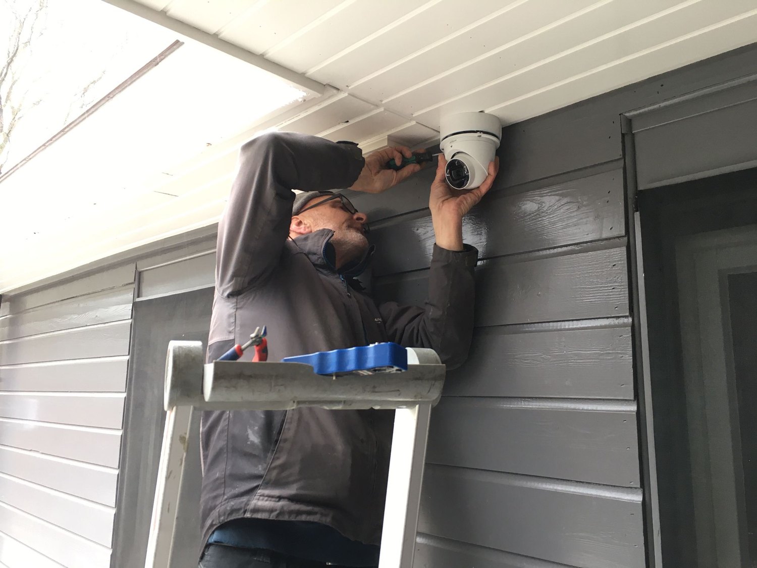 Monteur op een trap bevestigt een witte camera onder het witte overstek van een grijze houten gevel