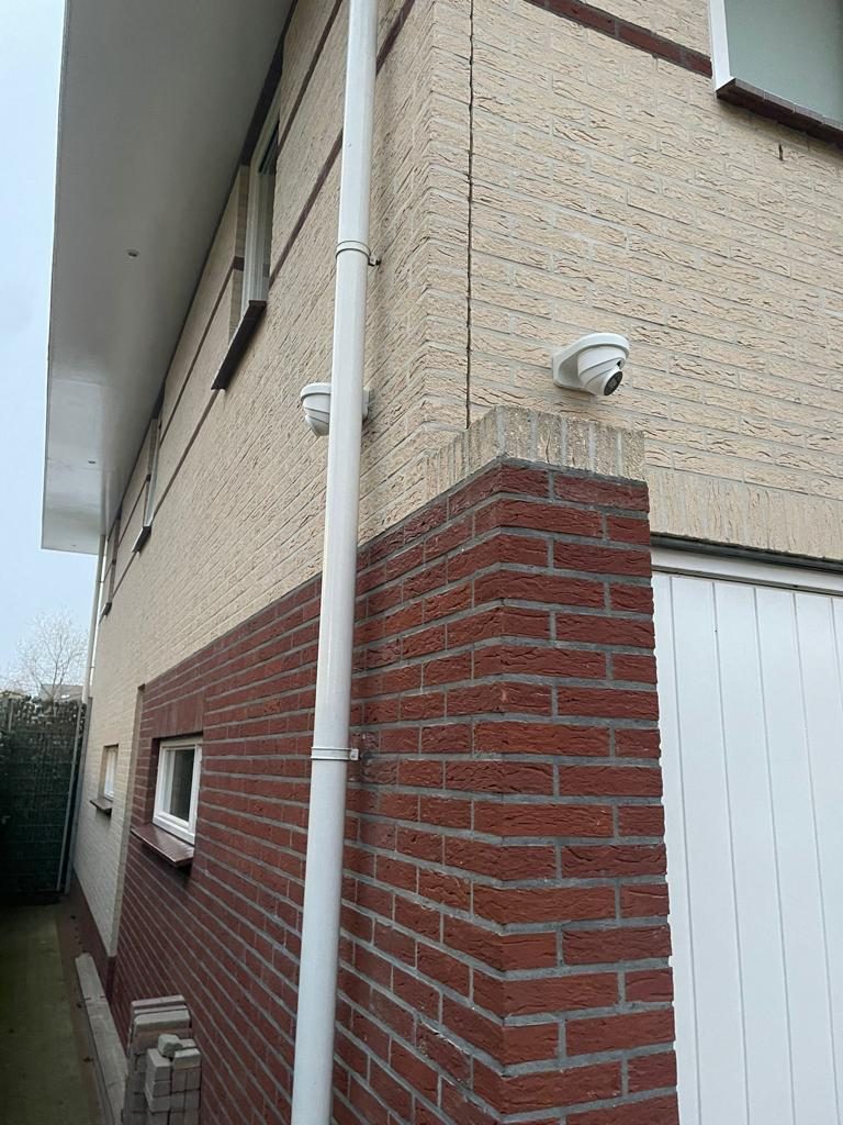 Twee witte camera's op de hoek van een woning: één gericht op de oprit, één op het achterompad langs de garage
