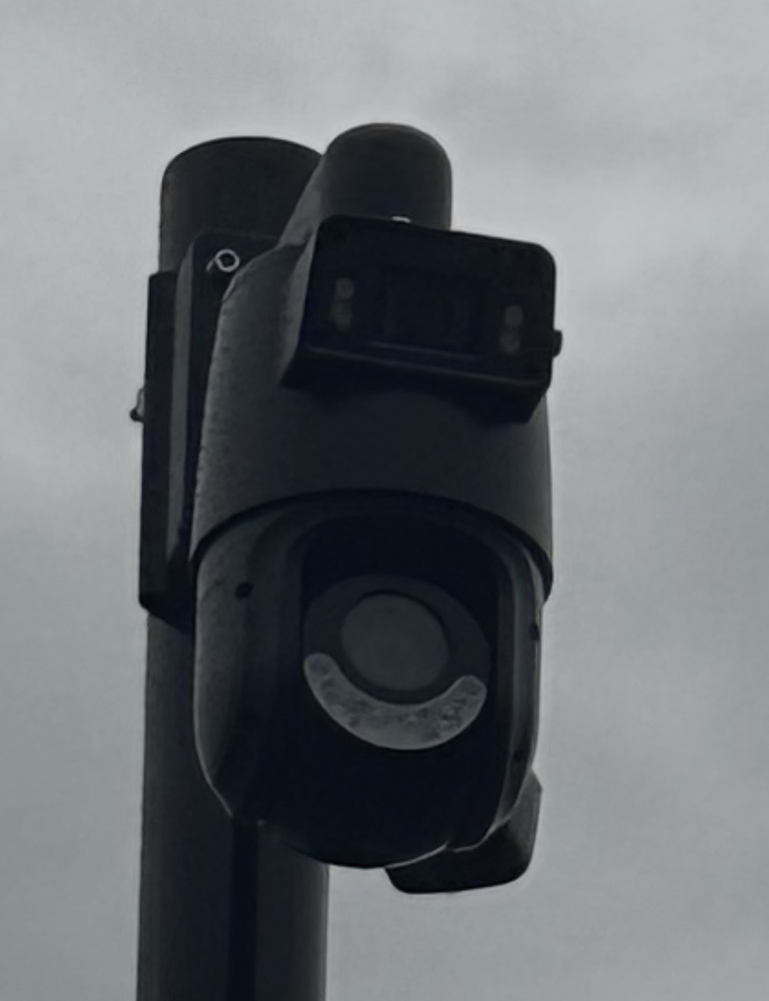 Zwarte draai- en kantelcamera met een eigen schijnwerper erboven, gemonteerd op een donkere mast tegen een grauwe lucht