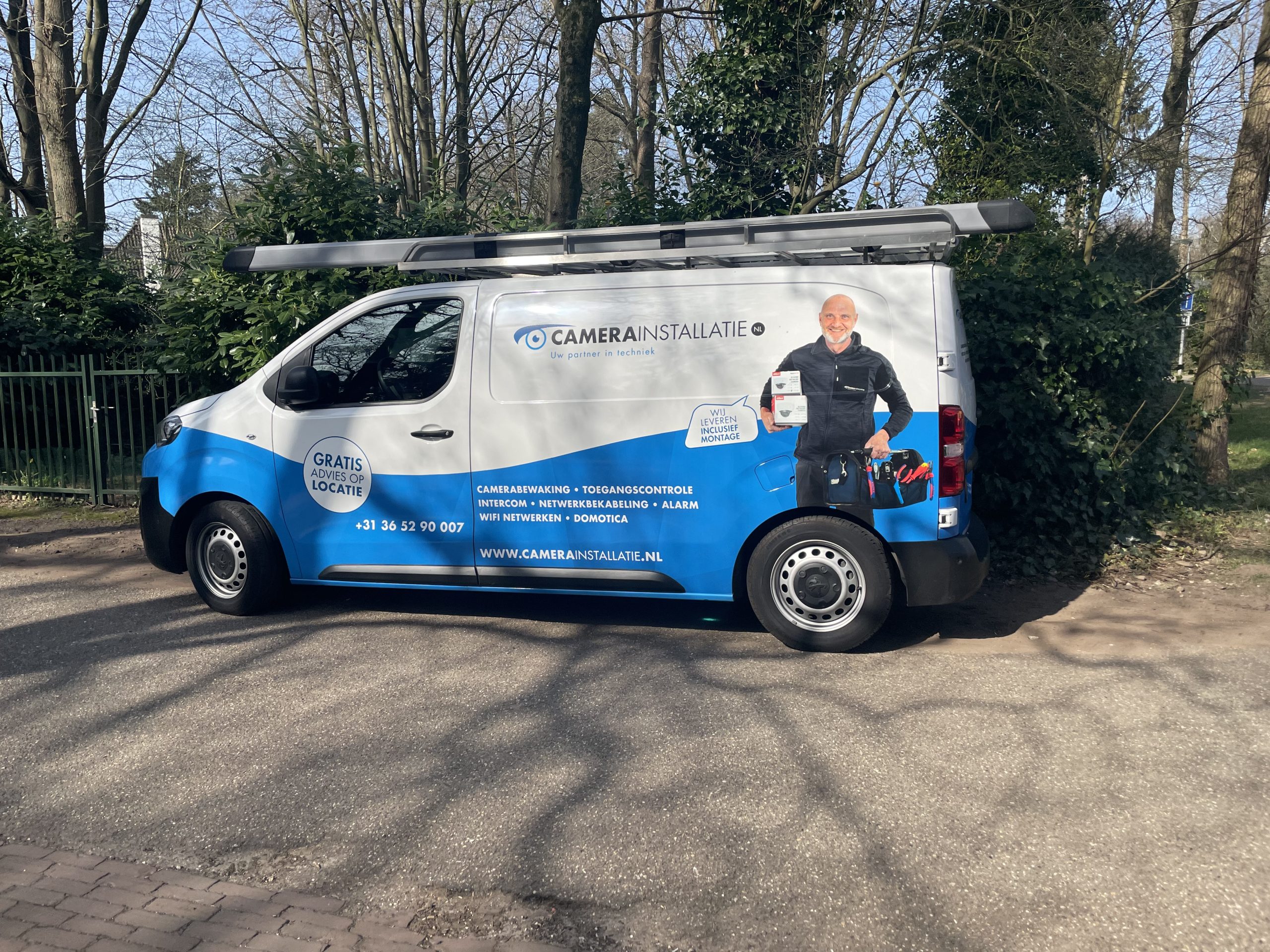 Servicebus van ons moederbedrijf CameraInstallatie.nl, geparkeerd langs de heg van een villa in een bosrijke straat