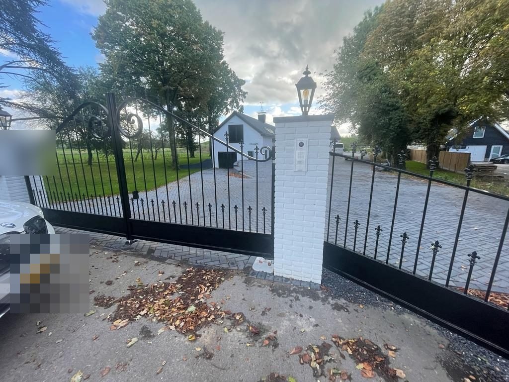 Smeedijzeren poort met intercom en codepaneel in een witte gemetselde pijler, met het landhuis op de achtergrond