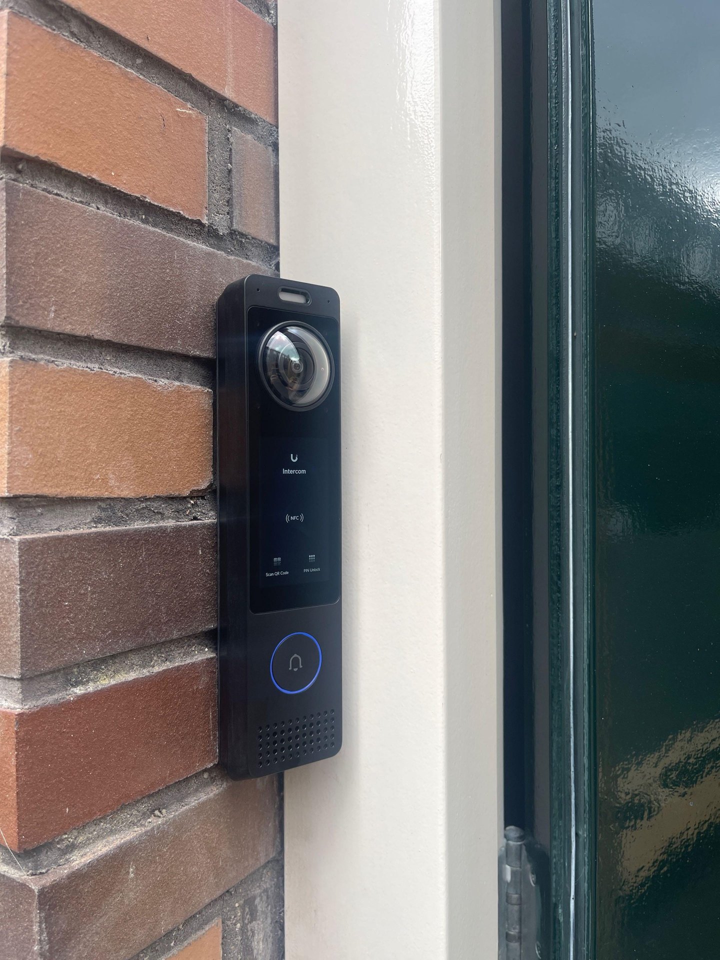 Zwarte video-intercom met pincode en QR-functie op een gemetselde muur naast een donkergroene deur