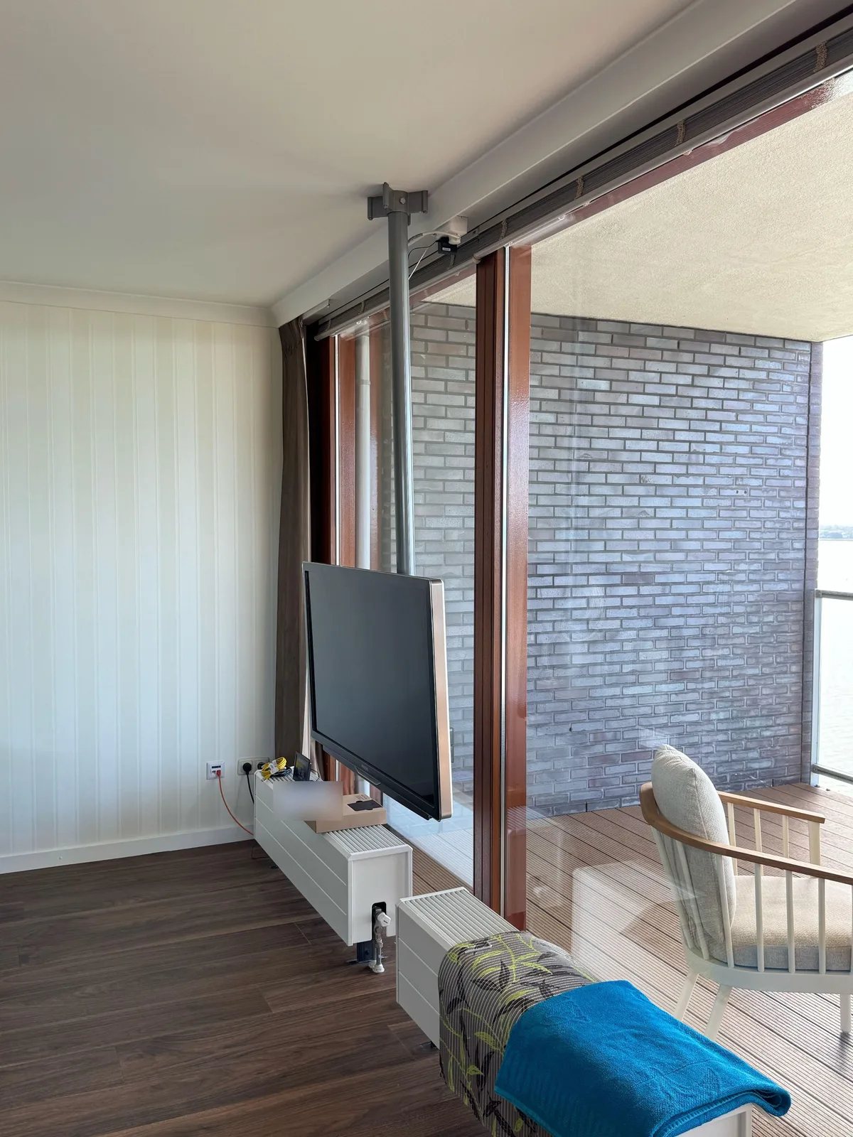 Lichte woonkamer met een tv aan een paal vanaf het plafond, naast een groot raam met balkon