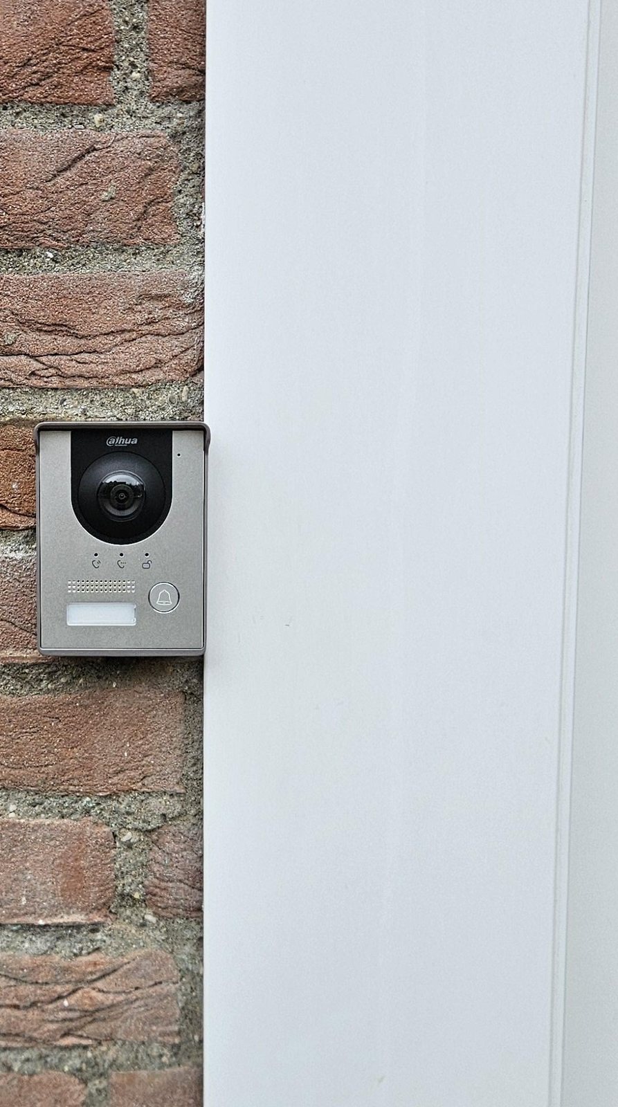 Buitenpost van een intercom met regenkap, naast een witte deurlijst