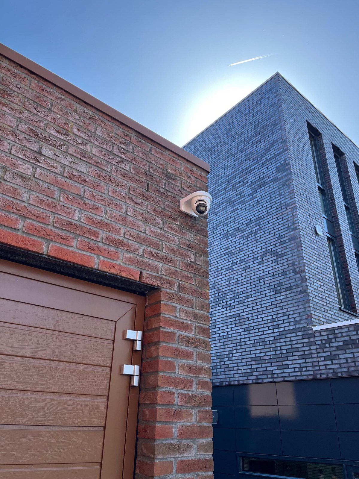 Witte camera op de hoek van een bakstenen muur boven een houten garagedeur, met een moderne gevel erachter