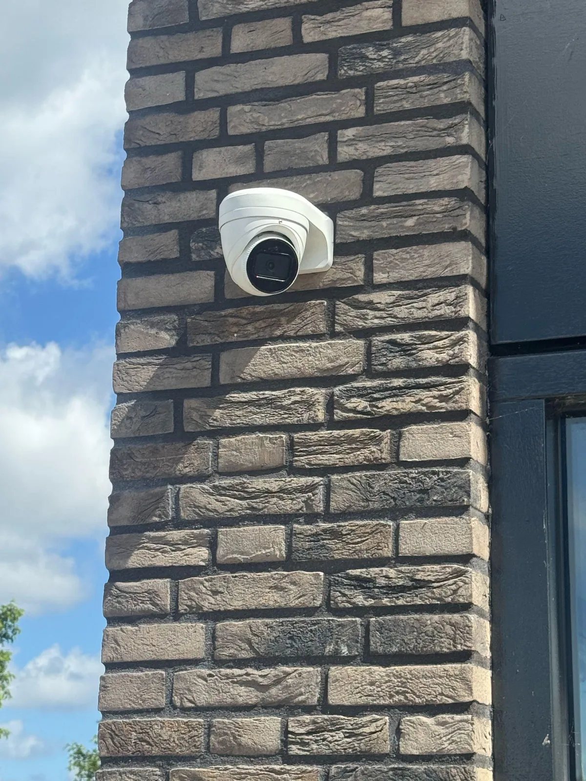 Witte turretcamera op een muurbeugel tegen donkere baksteen, naast een zwart raamkozijn