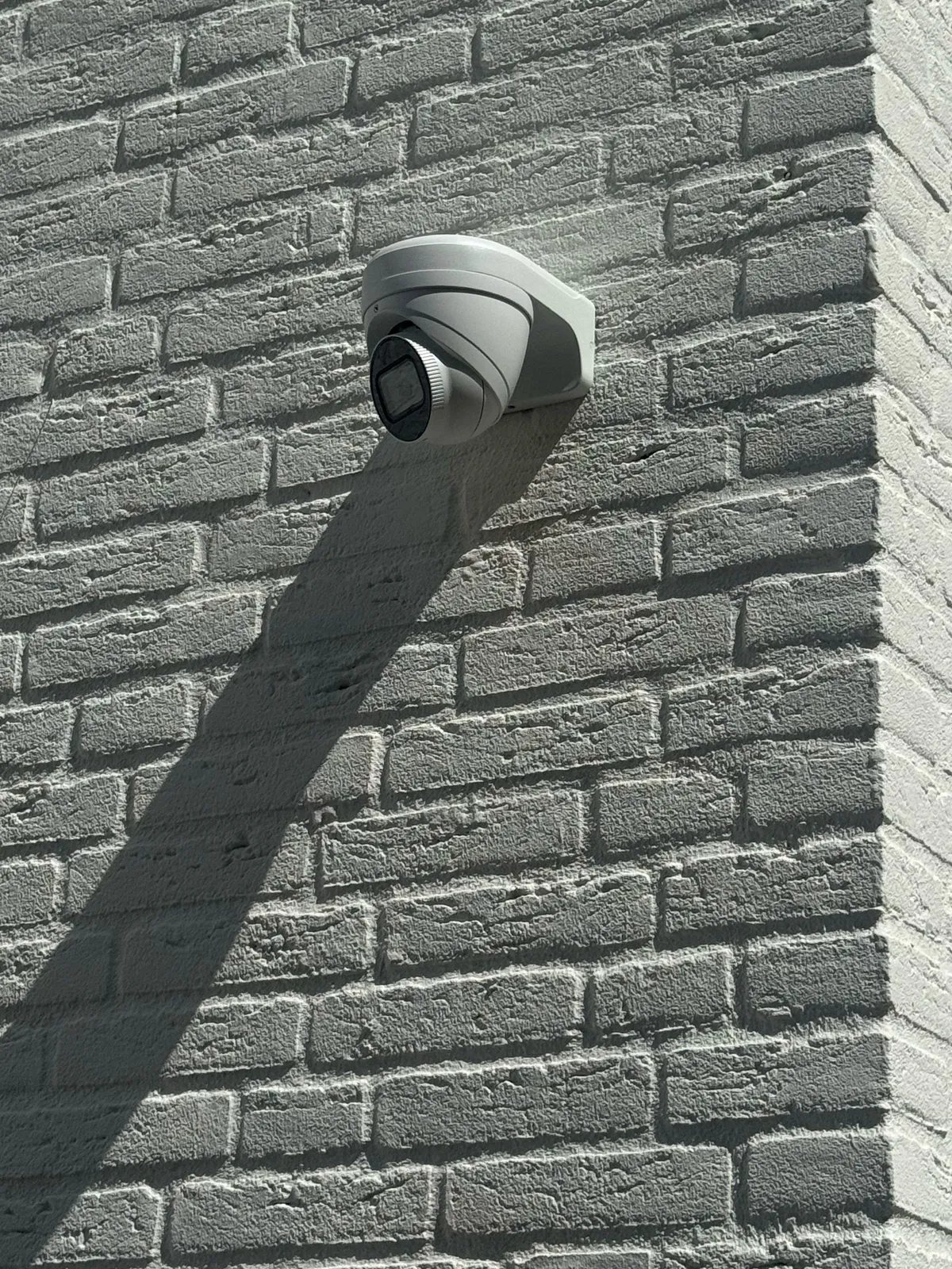Witte turretcamera op een muurbeugel op een lichte bakstenen gevel met een scherpe schaduw