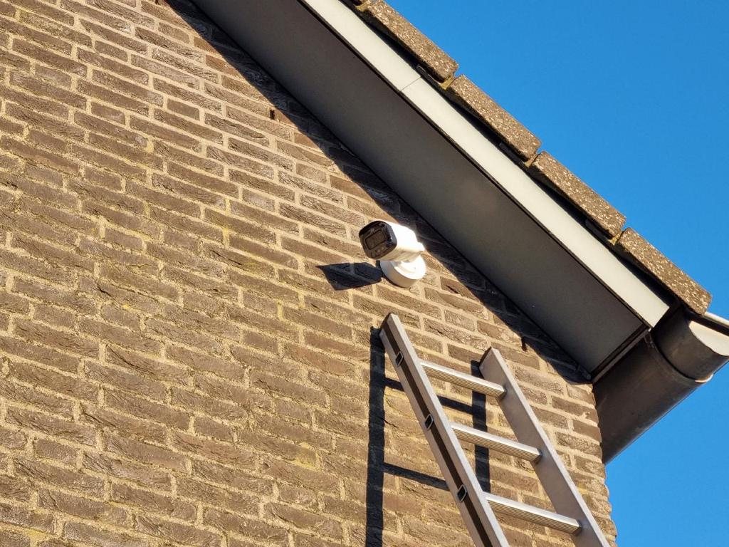 Witte camera hoog op een bakstenen gevel vlak onder de dakgoot, met een ladder ernaast