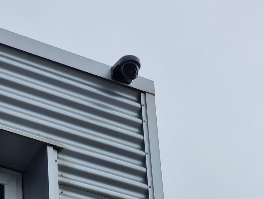 Zwarte camera op de bovenhoek van een bedrijfspand met grijze golfplaat, tegen een grijze lucht