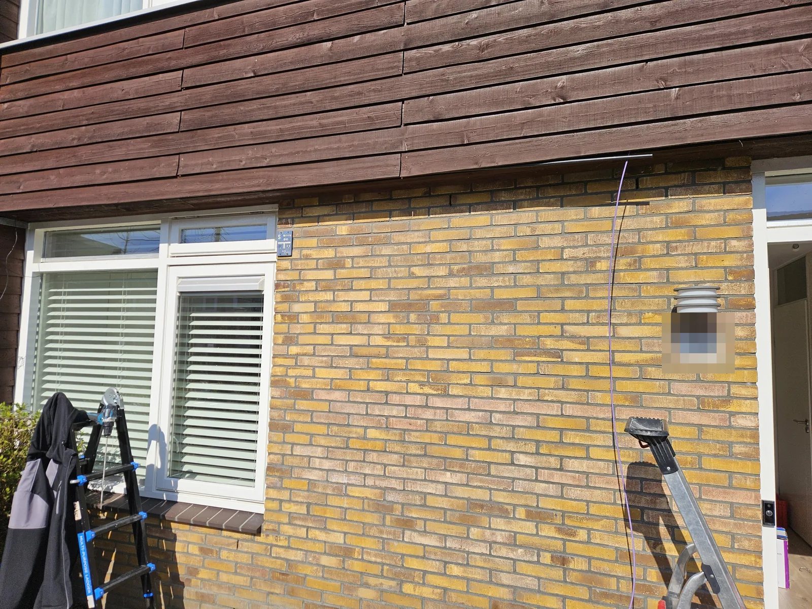 Gevel van gele baksteen met ladders, een paarse kabel hangt langs de muur en verdwijnt onder de houten betimmering