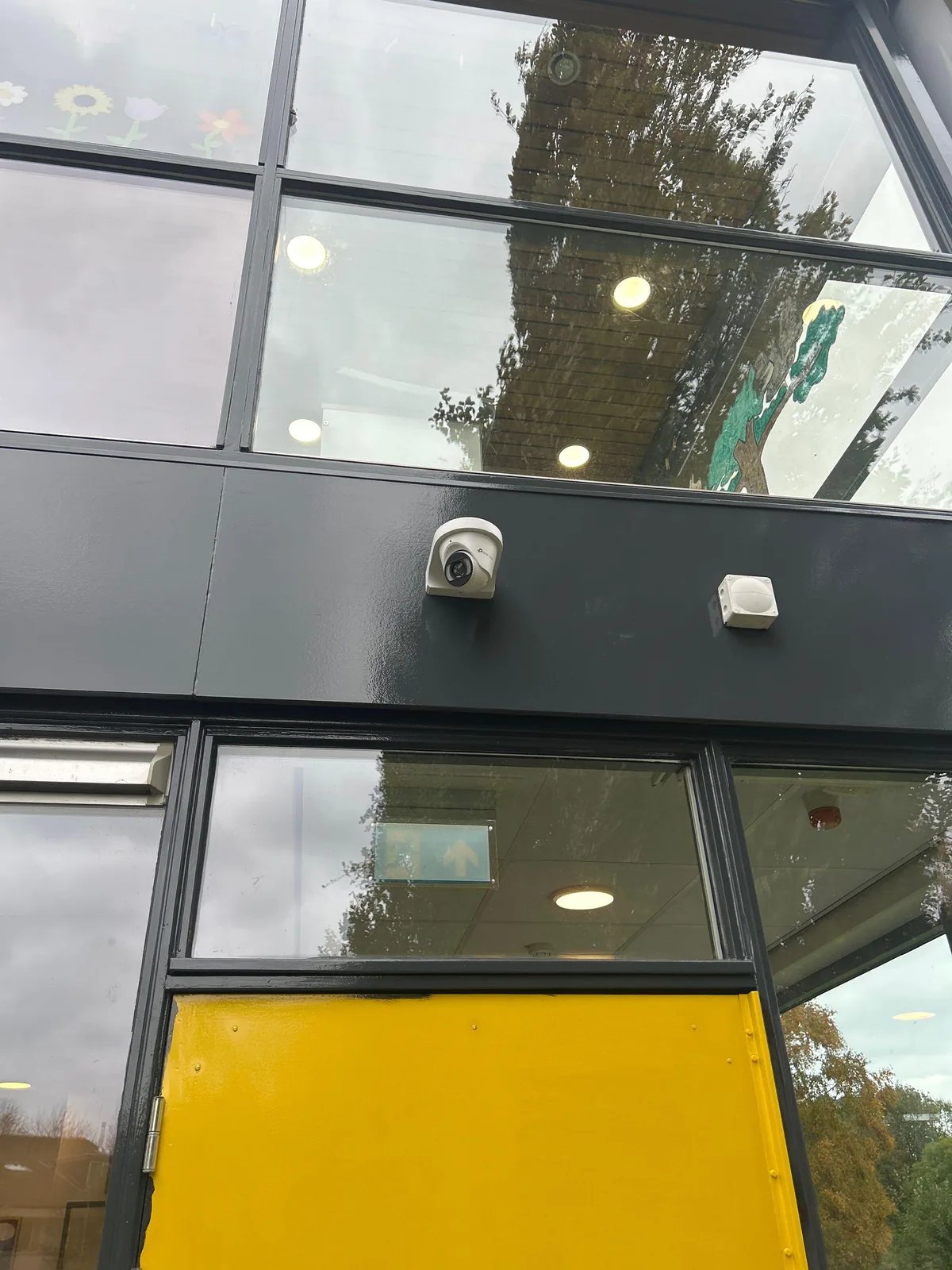 Witte camera op een donkere gevelband boven een gele deur van een kinderdagverblijf