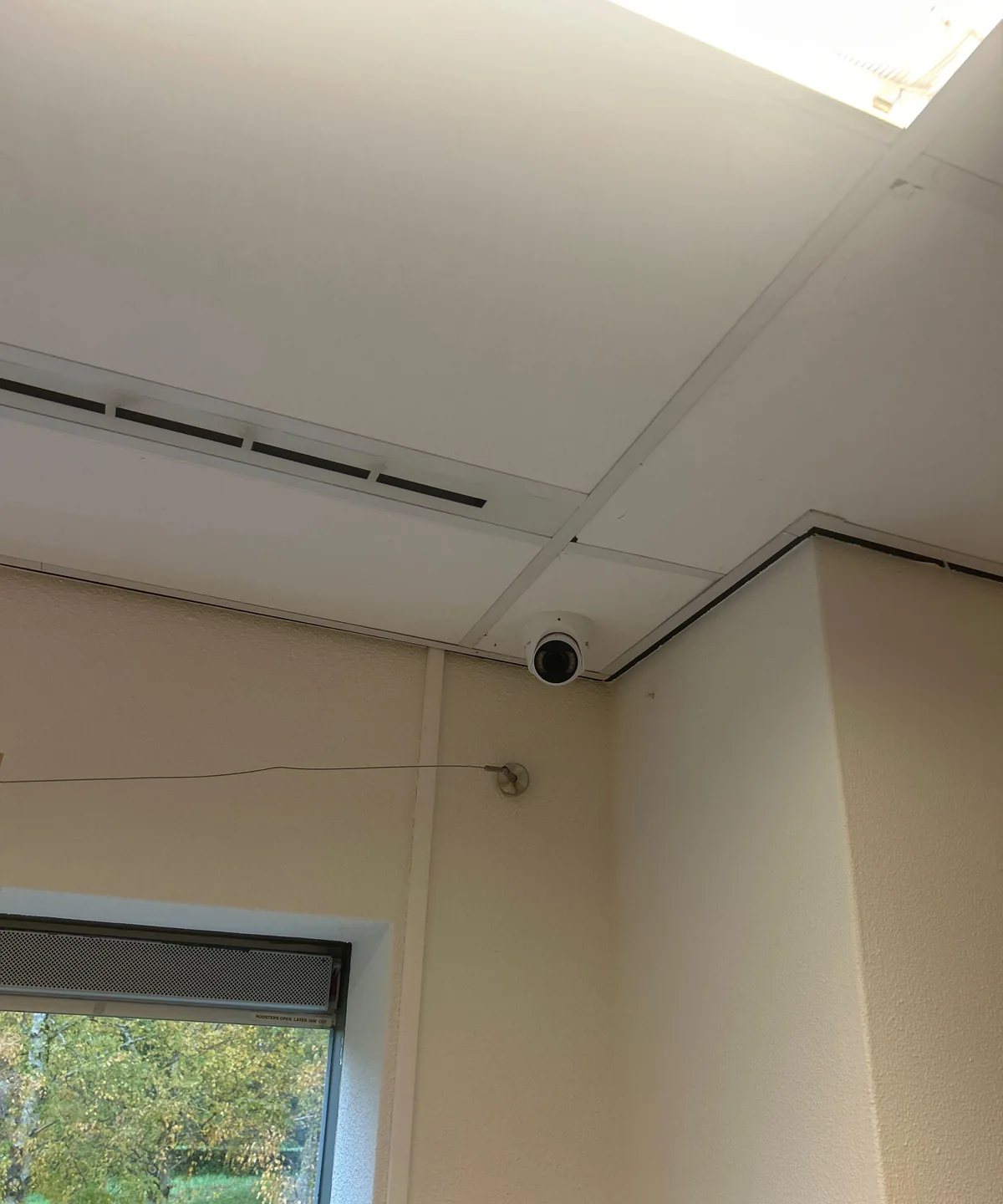 Witte camera hoog in de hoek van een groepsruimte tegen het systeemplafond, naast een raam met bomen erachter