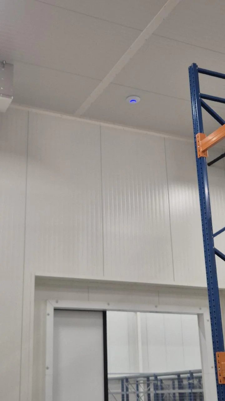 Rond access point met blauw lampje aan het plafond van een koelcel, boven een schuifdeur en een stelling