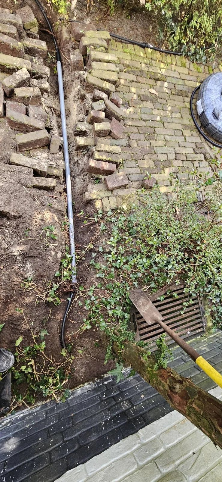 Open sleuf in een tuin met een grijze buis die onder een bakstenen looppad door gaat