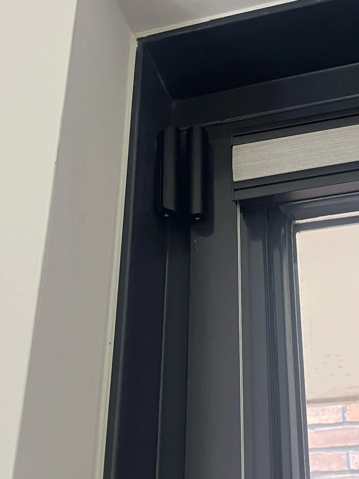 Zwart deurcontact boven in de hoek van een zwart kozijn, naast een rolgordijn