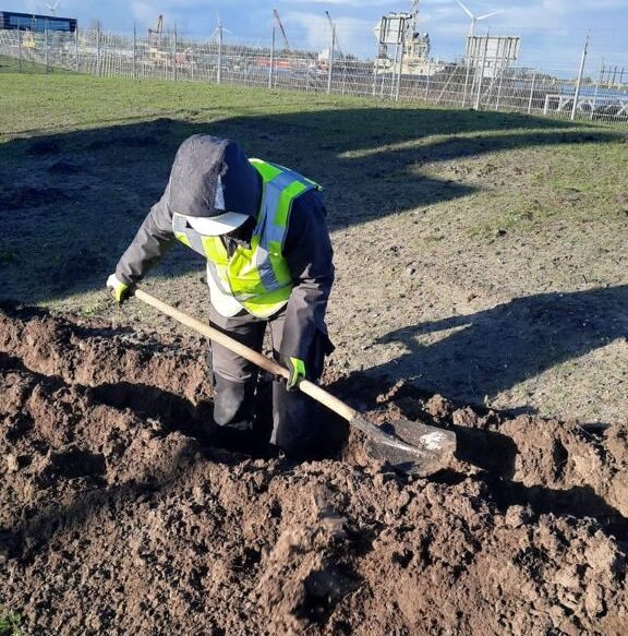 Monteur in geel hesje graaft met een schop in natte grond op een open terrein
