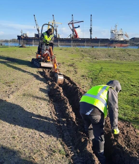 Minigraver trekt een sleuf door het gras, een monteur in geel hesje ernaast, op de achtergrond een schip en kranen