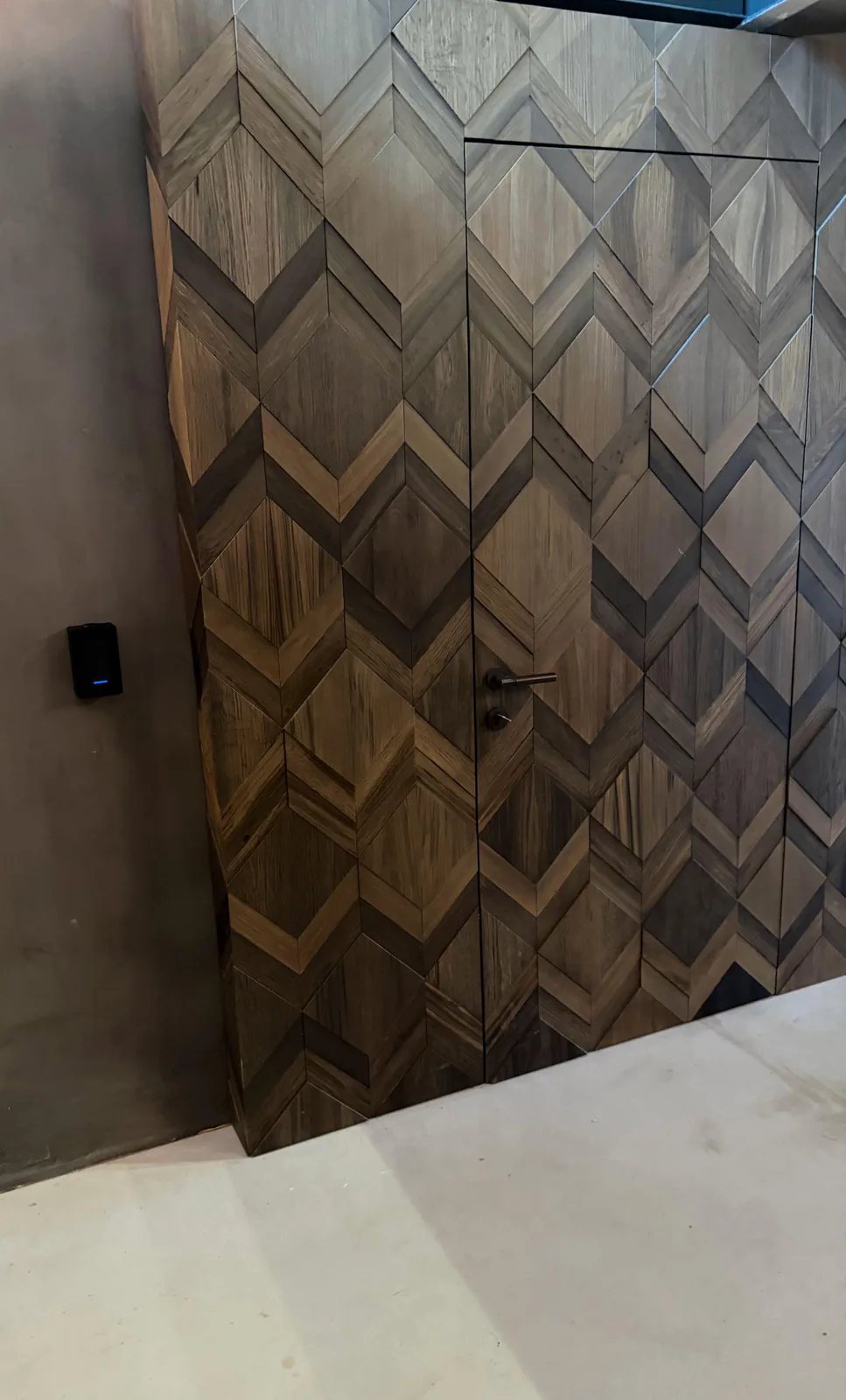 Zwarte toegangslezer met blauw lampje op een grijze wand, naast een deur die bijna verdwijnt in houten ruitpanelen