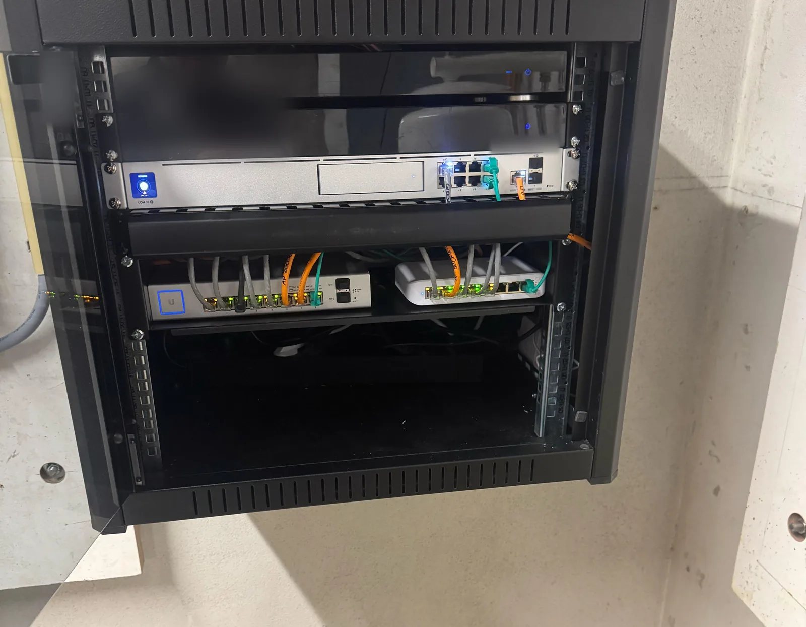 Open zwarte patchkast aan een betonwand met een UniFi-router en twee switches met grijze en oranje kabels