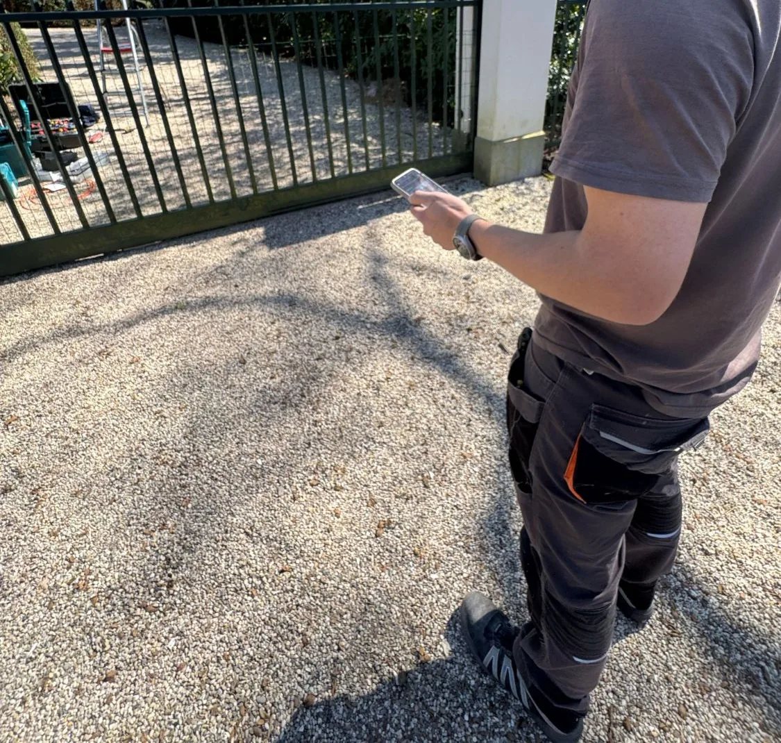 Monteur van achteren gezien met een telefoon in de hand voor een groene schuifpoort op een grindoprit