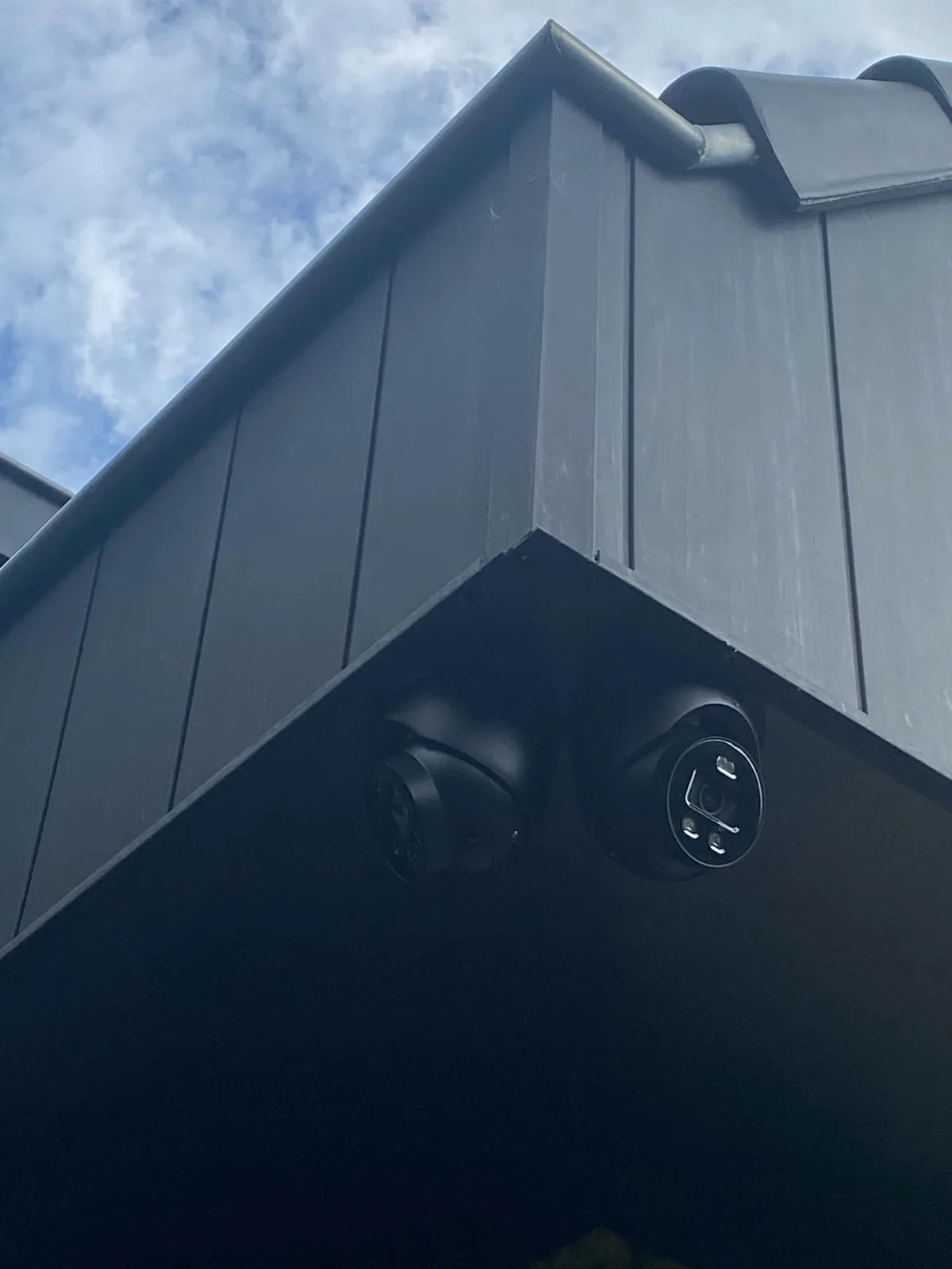 Twee zwarte camera's naast elkaar in de hoek onder een donker overstek, tegen een bewolkte lucht