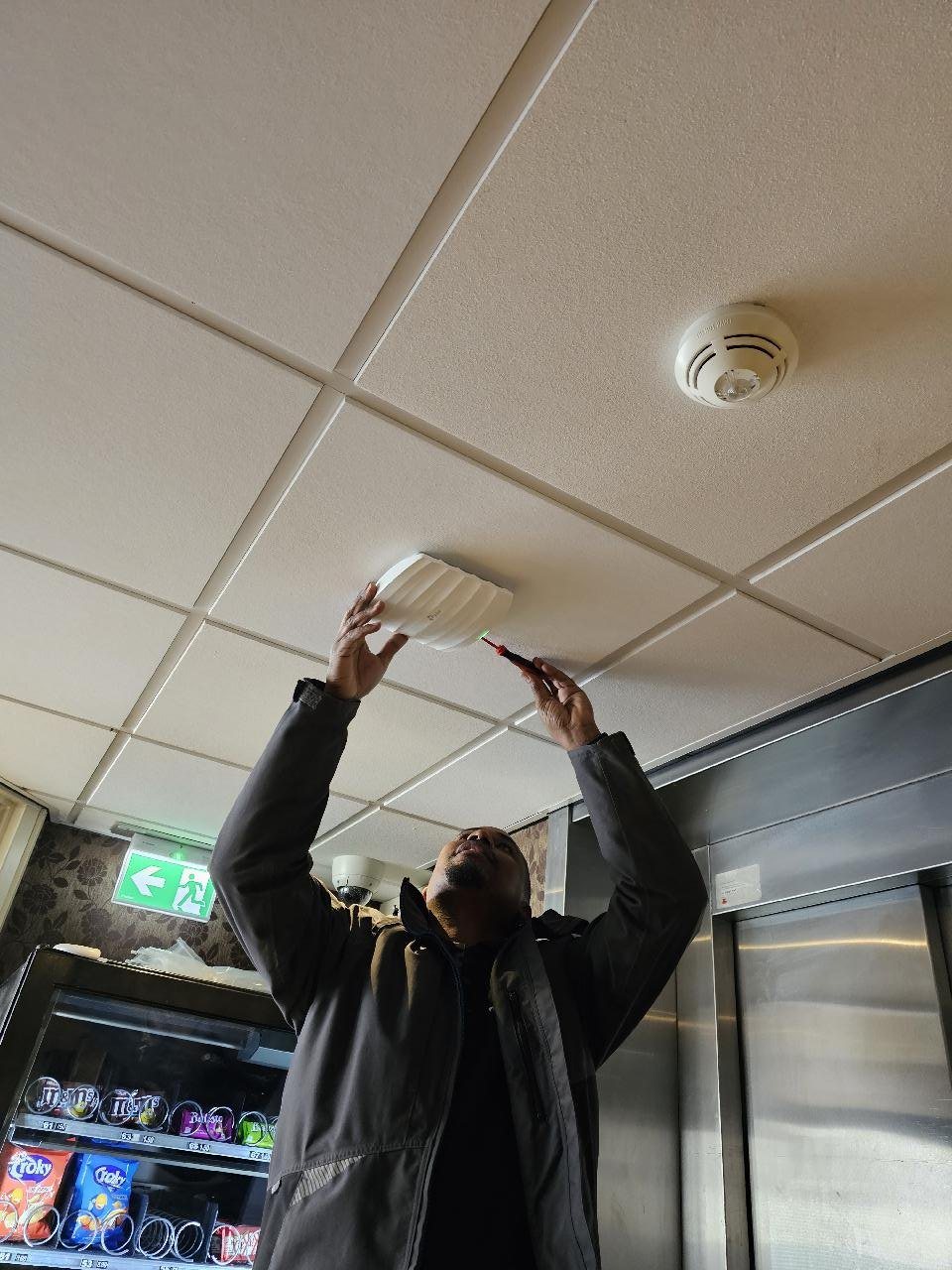 Monteur op een trapje hangt een witte wifizender aan het plafond van een hotellobby, naast een rookmelder