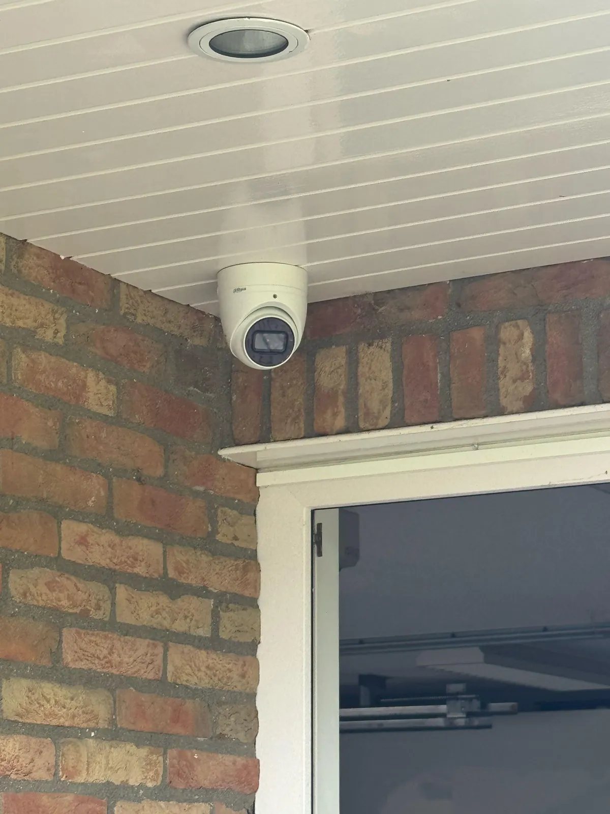 Witte turretcamera tegen een wit overstek met schroten, boven een deur met wit kozijn in rode baksteen