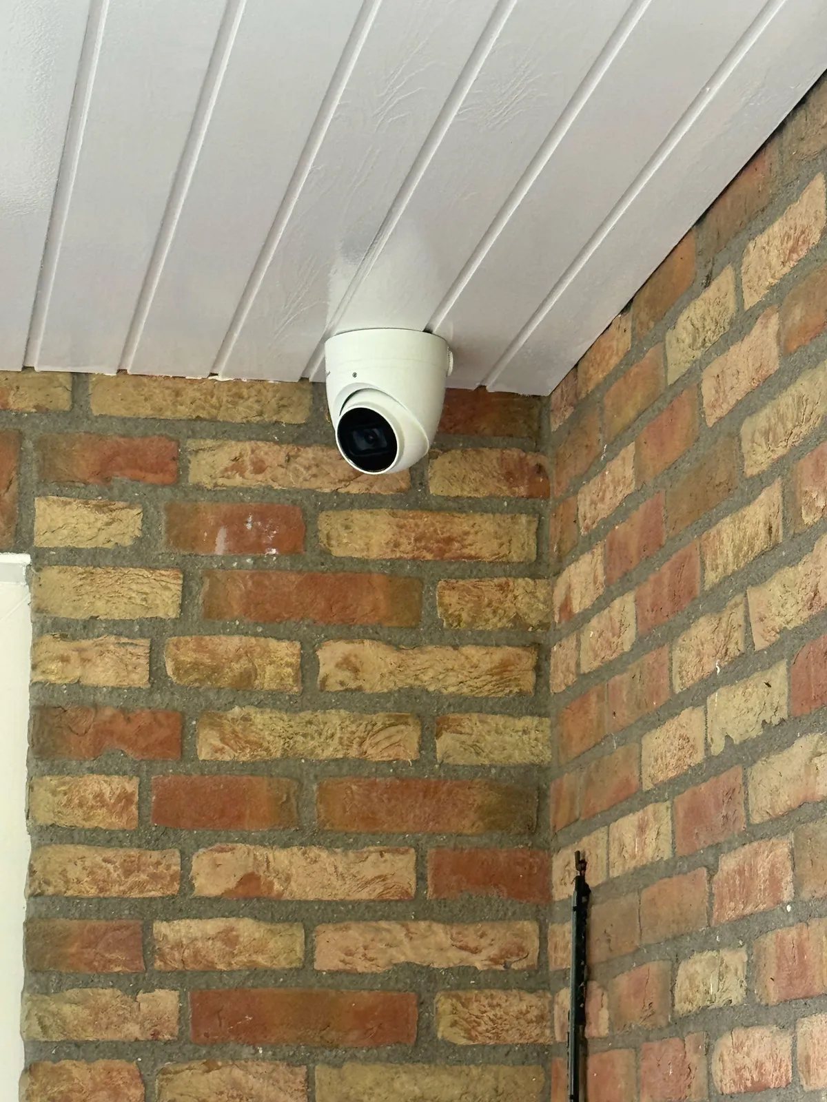 Witte turretcamera in de hoek onder een overstek met witte schroten, tegen rode en gele baksteen