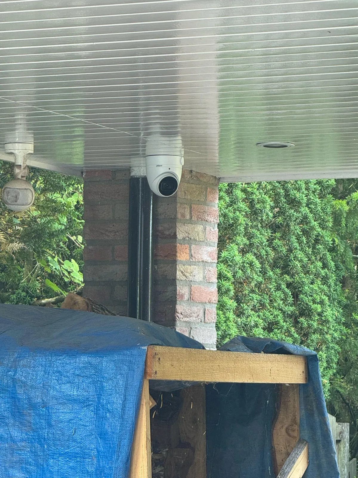 Witte camera onder een overkapping bij een bakstenen pilaar, met een groene haag erachter