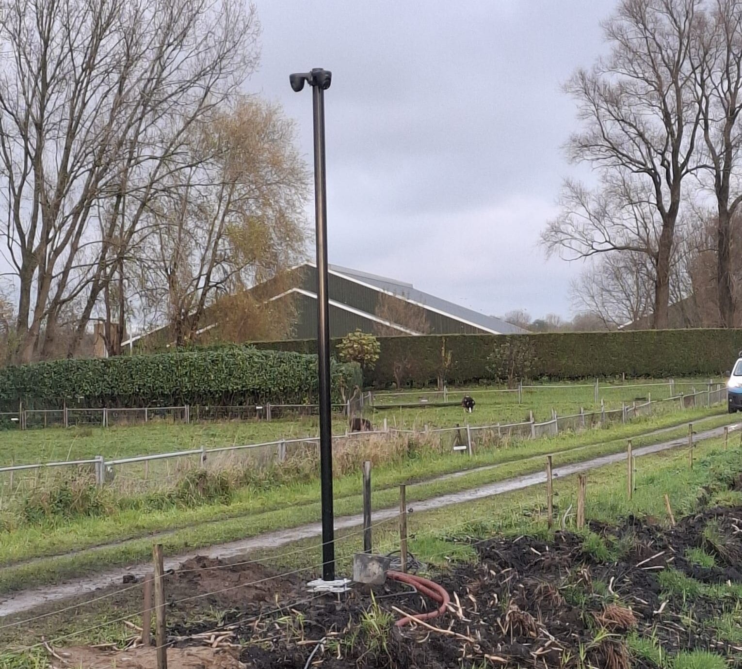 Camera op een donkere mast langs een haag, met een weiland erachter