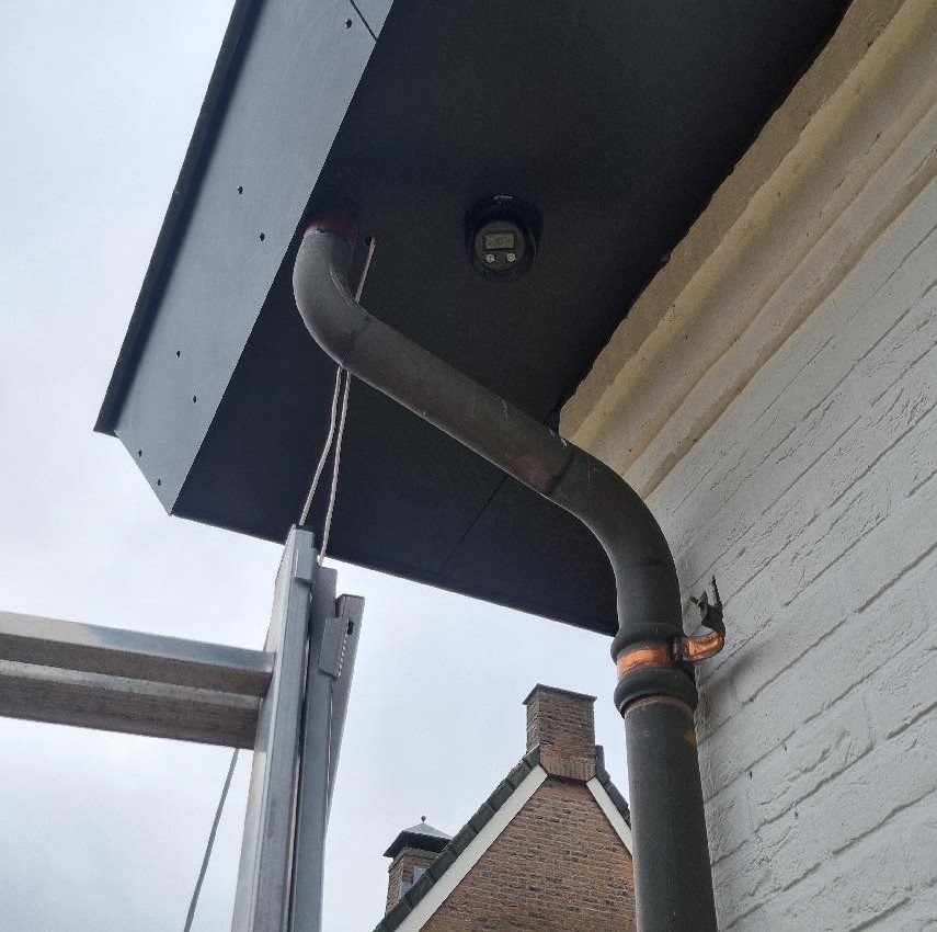 Zwarte camera in de onderkant van een zwarte dakrand, met een regenpijp ernaast