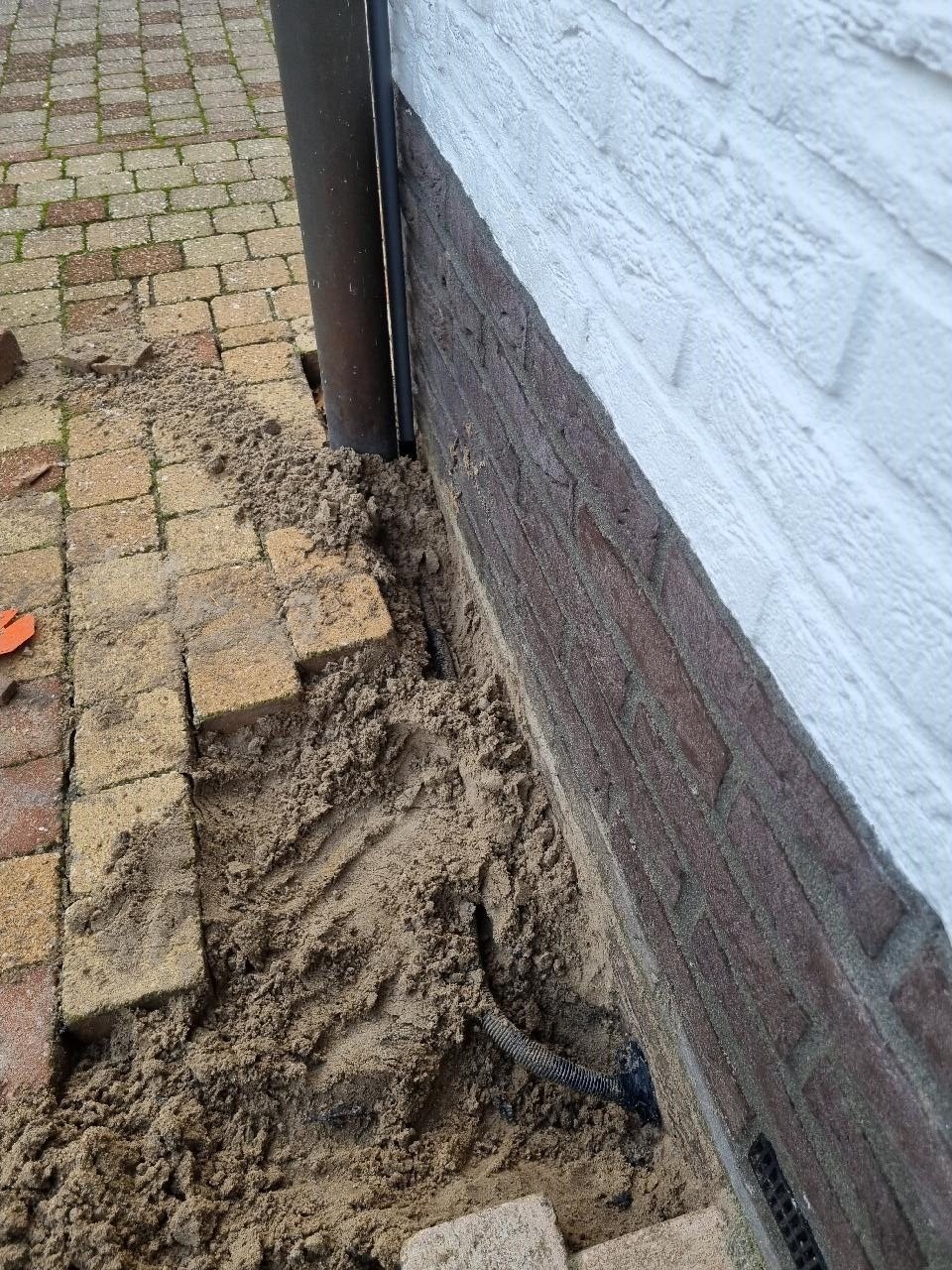 Open sleuf langs een witte gevel met een kabelbuis erin, tegels opzij gelegd